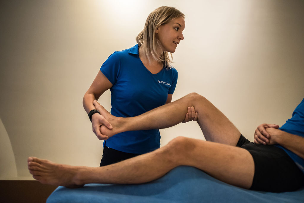 Fysiotherapeut Brigitte in een blauw shirt helpt een patiënt aan zijn liesblessure en hamstring die op de behandelbank ligt.