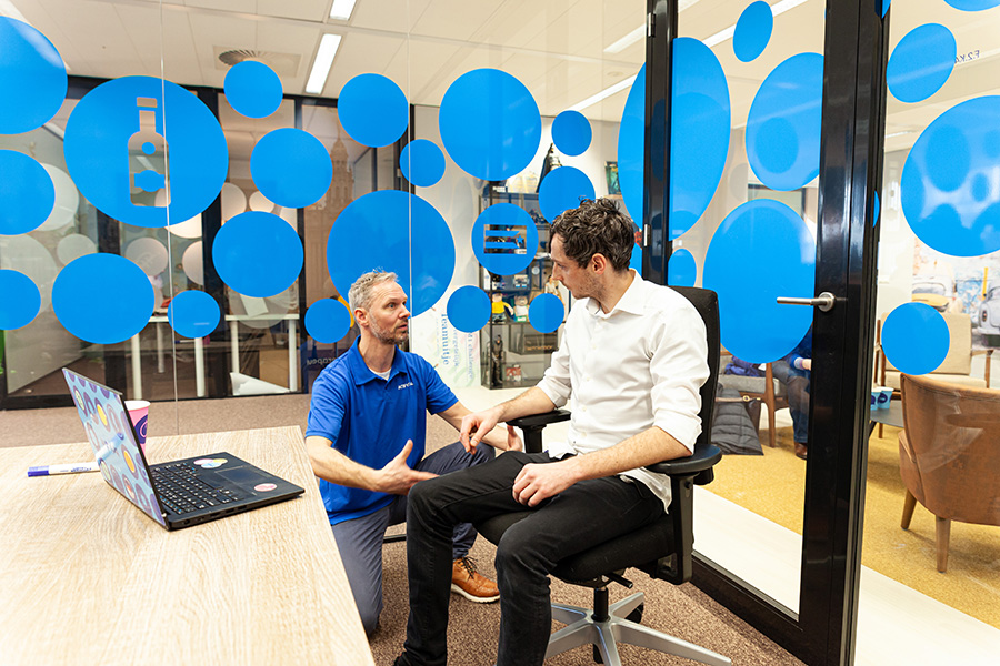 Fysiotherapeut Robert Lutz in een blauw jasje legt aan een medewerker van Bol.com uit over de juiste zithouding via het bedrijfsaanbod.