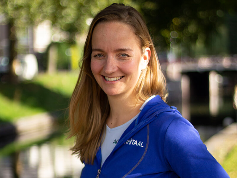 Psychosomatisch fysiotherapeut Laura Gilhuis poseert voor de camera in een wit shirt en blauwe hoodie op een zonnige dag.