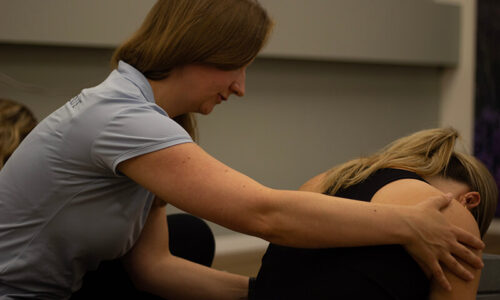 Fysiotherapeut in een grijze polo geeft Yin Yoga aan een patiënt in de oefenzaal.