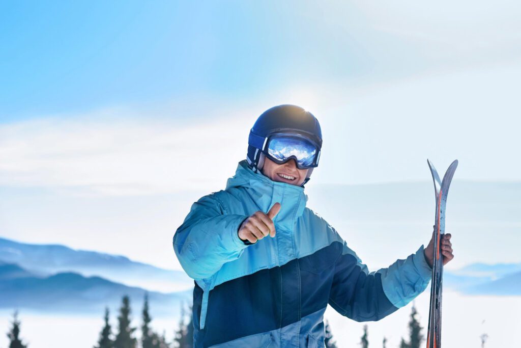 Een man houdt in zijn ene hand zijn ski's vast en met zijn andere hand steekt hij zijn skiduim op.