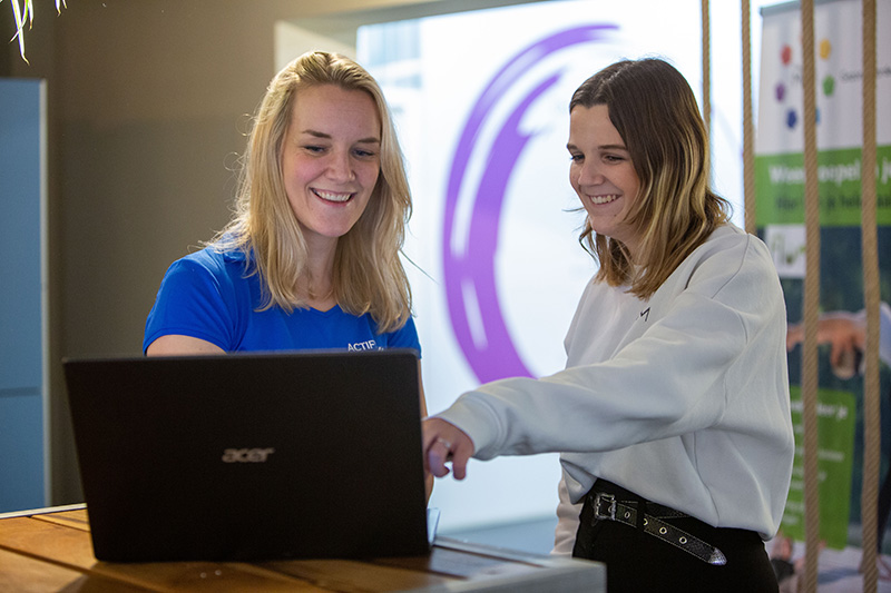 Patiënt in een witte sweater komt opdagen voor haar eerste fysiotherapie afspraak en wijst naar de laptop samen met de fysiotherapeut in een blauw shirt.