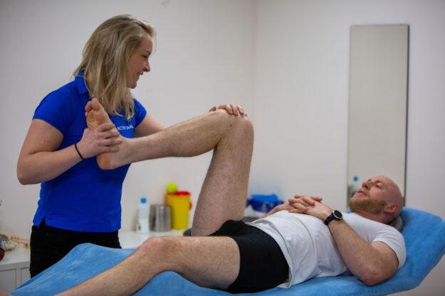 Man in een wit shirt wordt geholpen aan zijn knie door een fysiotherapeut in een blauw shirt op de behandelbank, omdat leeftijd invloed op blessures en de achillespees heeft een sportblessure aan de knie.