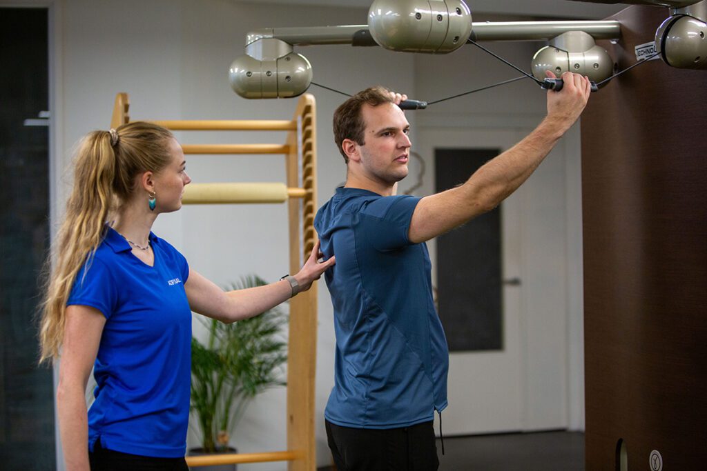 een man met een blauw shirt die begeleiding krijgt van de fysiotherapeute tijdens het uitvoeren van activatie bewegingen als krachtige warming-up bij een schouder uit de kom