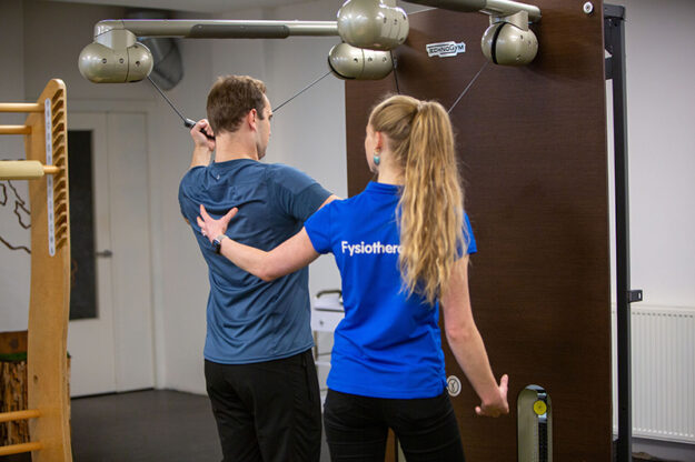 Man in een grijs shirt wordt geholpen door een fysiotherapeute in een blauw shirt in de oefenzaal en moet herstellen na het sporten.