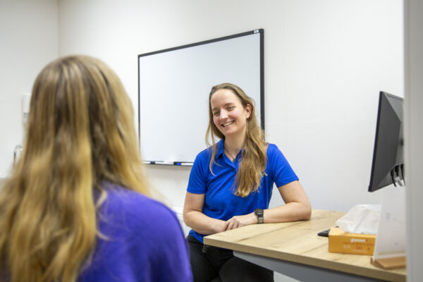 hielspoor Vrouwelijke patiënt in een paars shirt komt voor intake bij hielspoor bij de fysiotherapeut in een blauw shirt